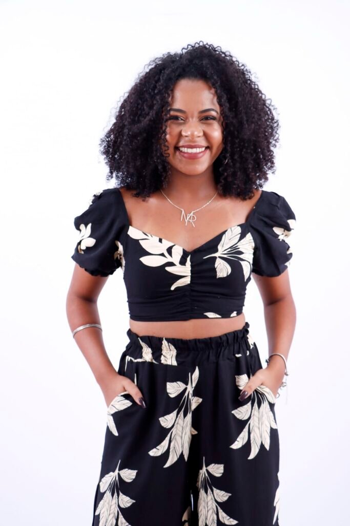 Cantora sorrindo com as mãos nos bolsos, de conjunto preto estampado, no fundo branco