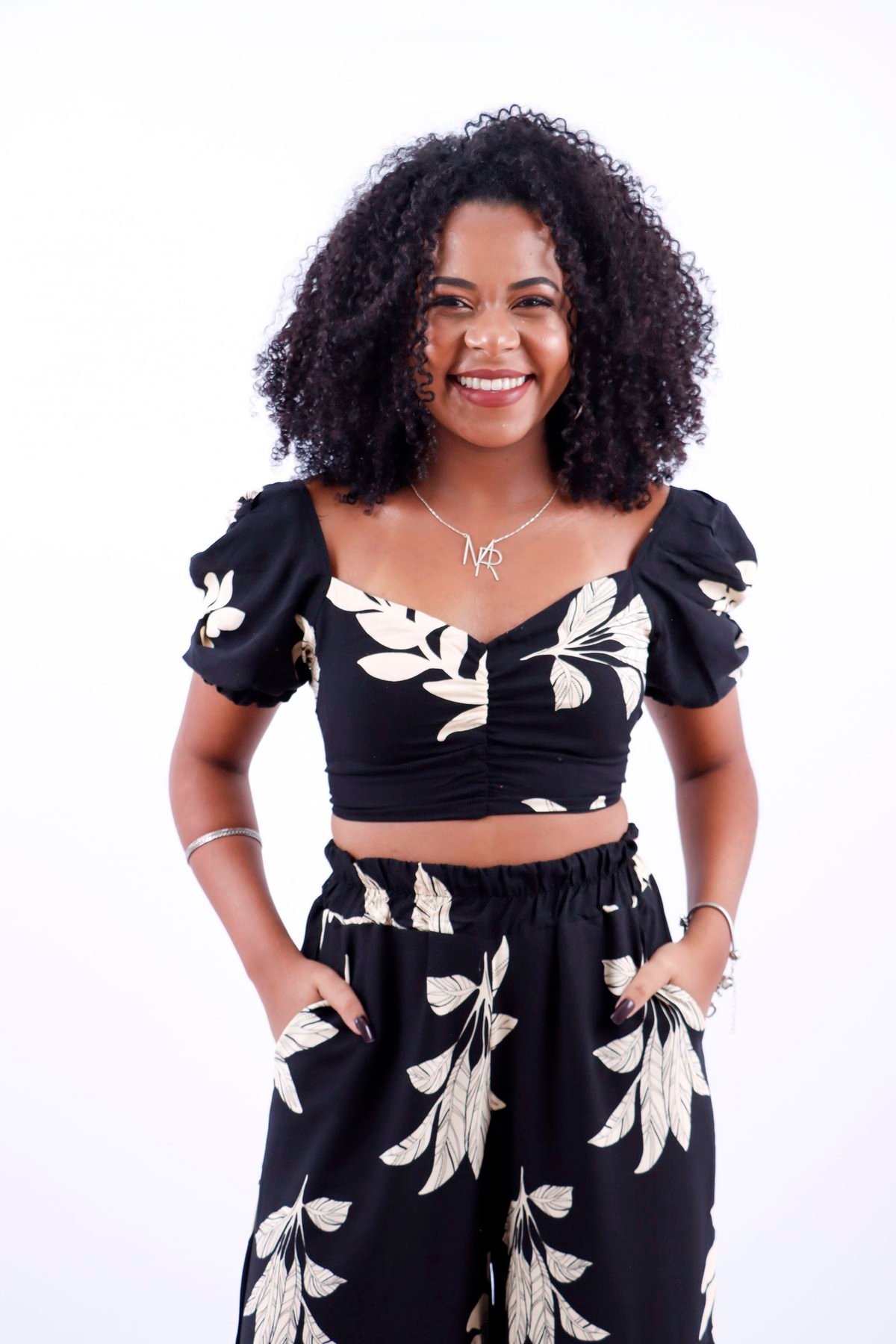 Cantora sorrindo com as mãos nos bolsos, de conjunto preto estampado, no fundo branco