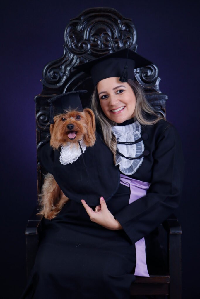Formanda de beca e faixa lilás na Cadeira Colonial Luiz XV com o cachorro yorkshire de beca e capelo no colo