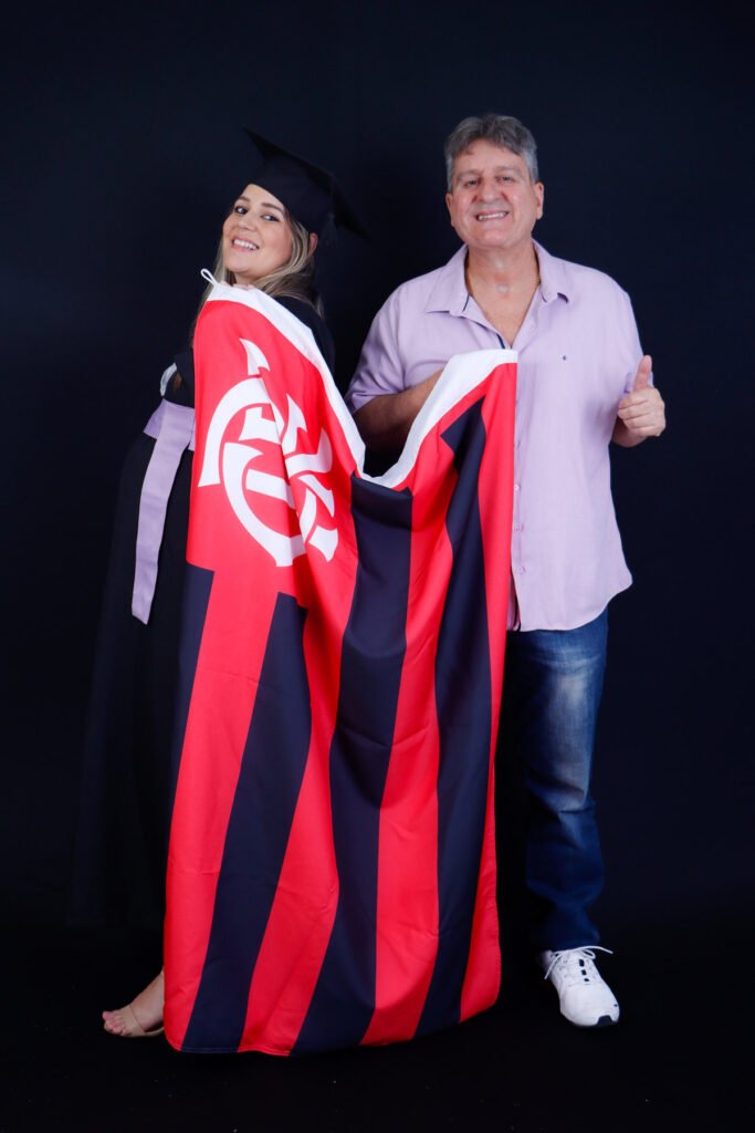 Formanda de beca enrolada na bandeira do time do coração ao lado de um familiar fazendo sinal de positivo