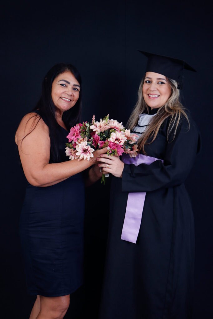 Formanda de beca e faixa lilás segurando um buquê de flores junto com uma familiar no fundo preto