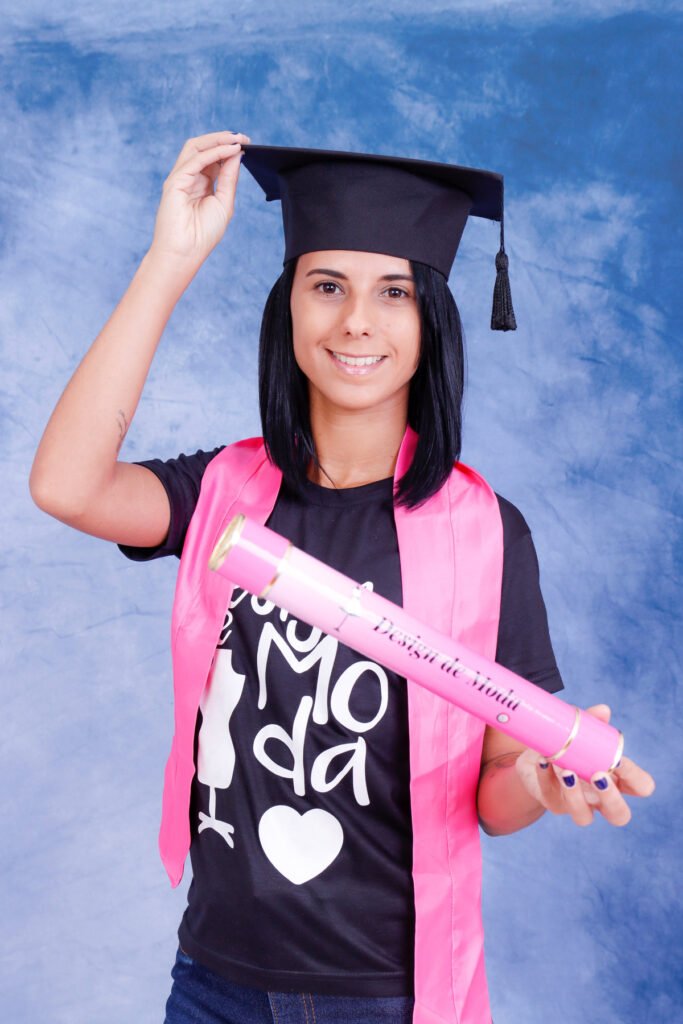Formanda de Design de Moda de capelo, camiseta do curso e faixa rosa segurando o canudo rosa no fundo azul