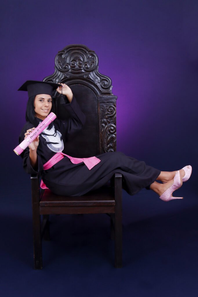 Formanda de Design de Moda de beca e faixa rosa sentada de lado na Cadeira Colonial Luiz XV, com sapatos rosa e o canudo do curso