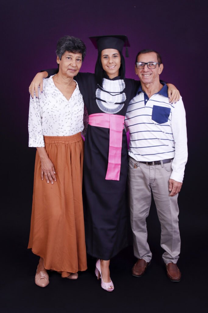 Formanda de Design de Moda de beca e faixa rosa abraçada a dois familiares no fundo roxo