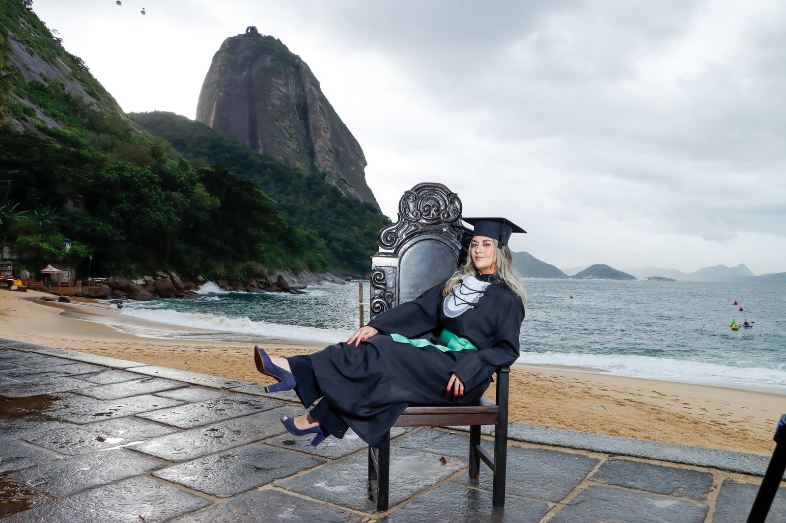 Formanda de Enfermagem de beca e faixa verde recostada na Cadeira Colonial Luiz XV no calçadão da Praia Vermelha, na Urca