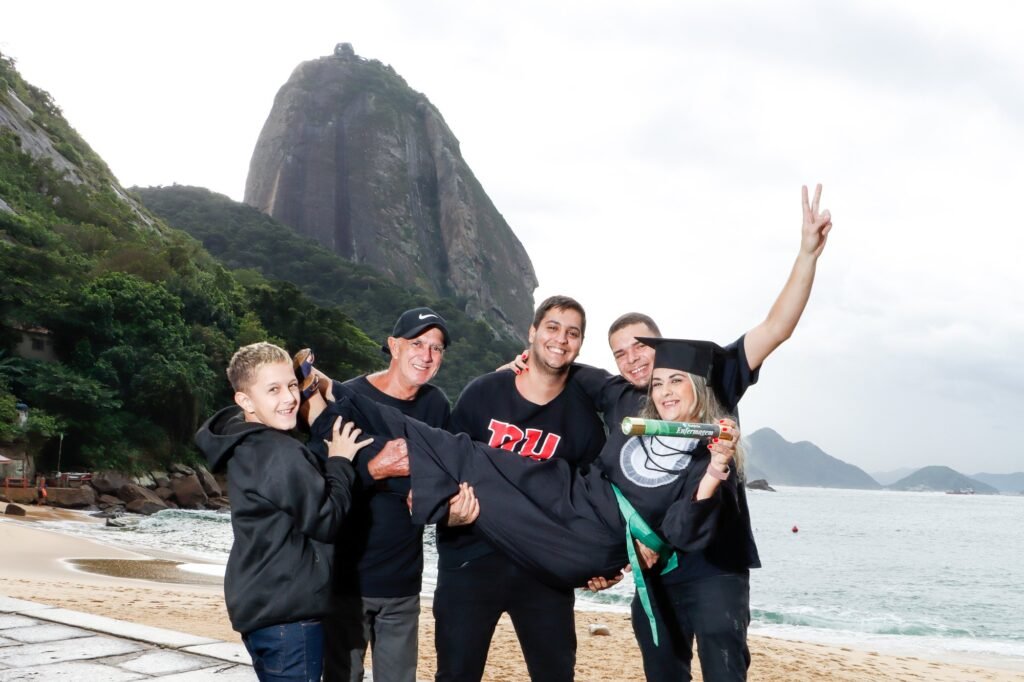 Formanda de Enfermagem carregada no colo pela família na areia da Praia Vermelha, segurando o canudo do curso