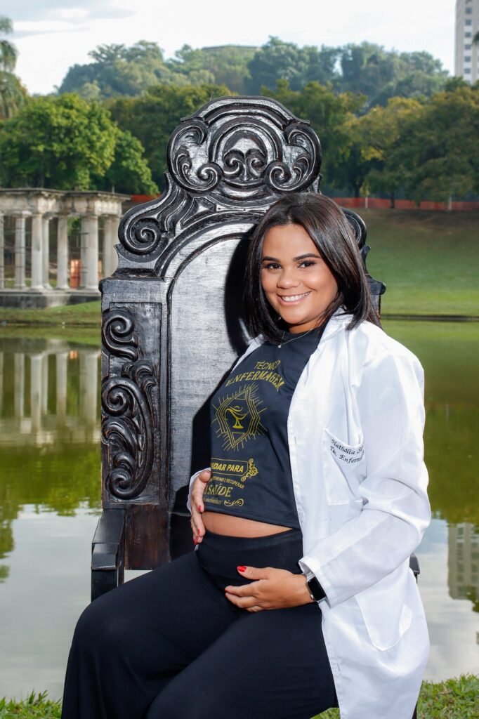 Formanda de Enfermagem de jaleco branco na Cadeira Colonial Luiz XV, com as mãos na barriga de gestante, no lago da Quinta da Boa Vista