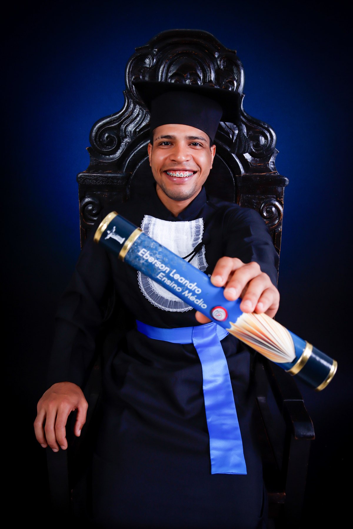 Formando do ensino médio de beca e faixa azul na Cadeira Colonial Luiz XV estendendo o canudo para a câmera