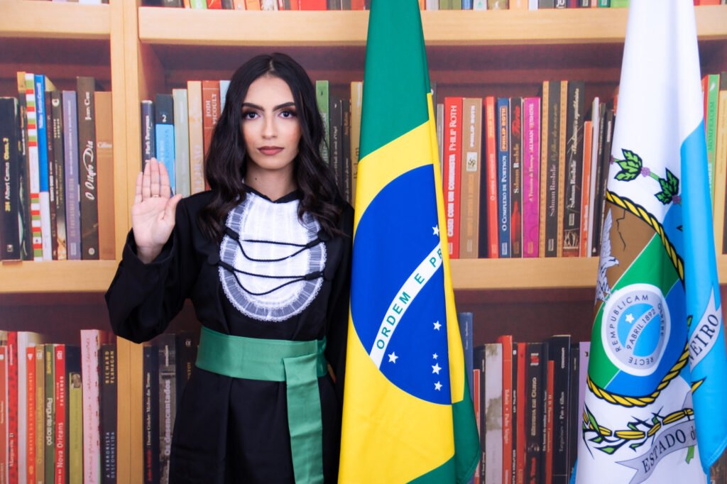 Formanda de beca com a mão erguida no juramento, ao lado das bandeiras do Brasil e do Rio de Janeiro