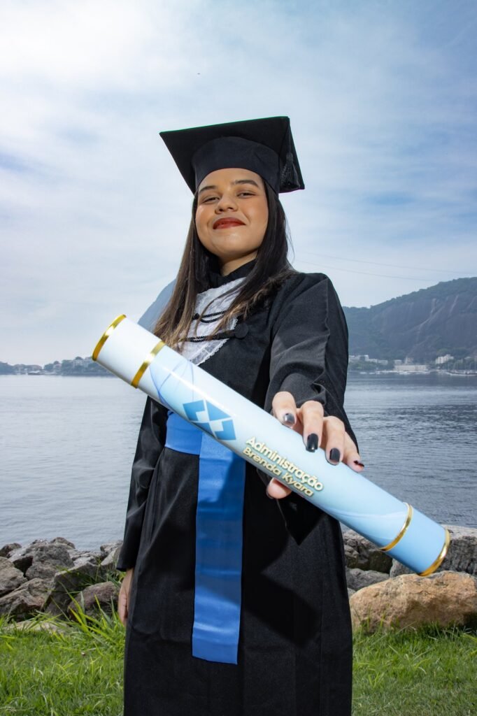 Formanda de Administração estendendo para a câmera o canudo personalizado com o nome do curso, à beira da baía