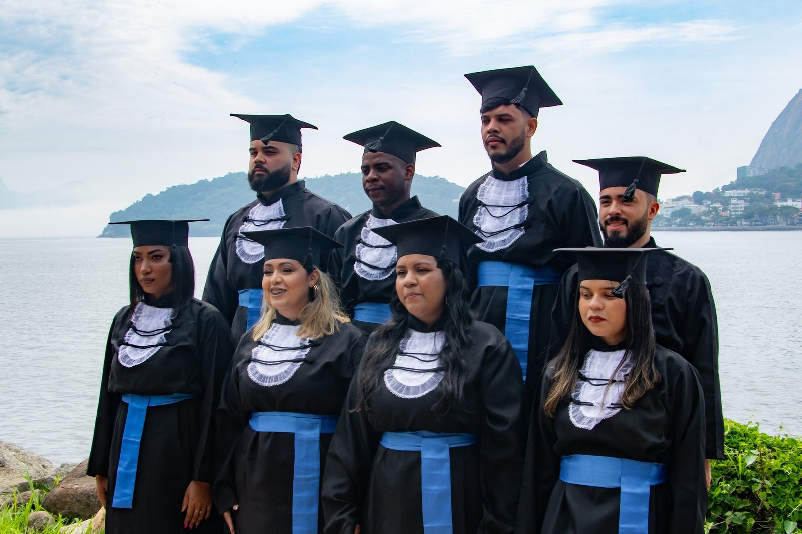 Turma de Administração de beca e faixa azul em duas fileiras à beira da baía, num dia nublado