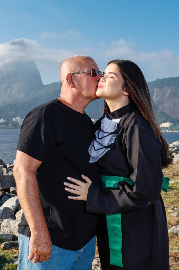 Formanda de beca e faixa verde recebendo um beijo no rosto do pai, à beira da baía