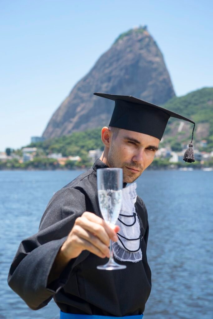 Formando de beca e capelo erguendo uma taça de espumante com o Pão de Açúcar ao fundo