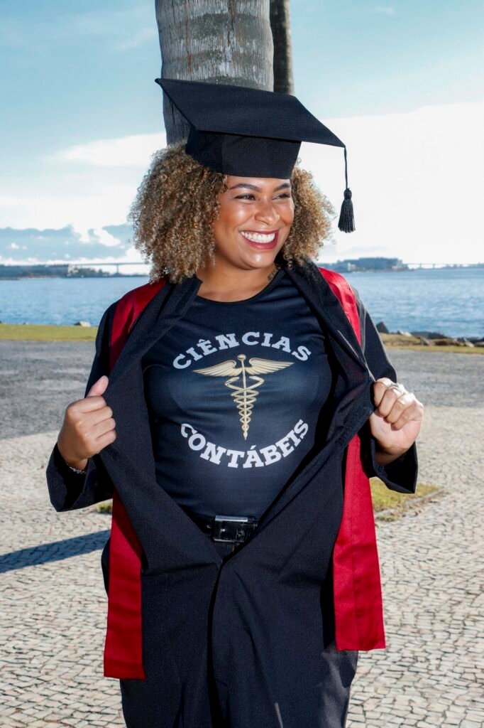 Formanda de Ciências Contábeis abrindo a beca para mostrar a camiseta do curso, à beira da baía