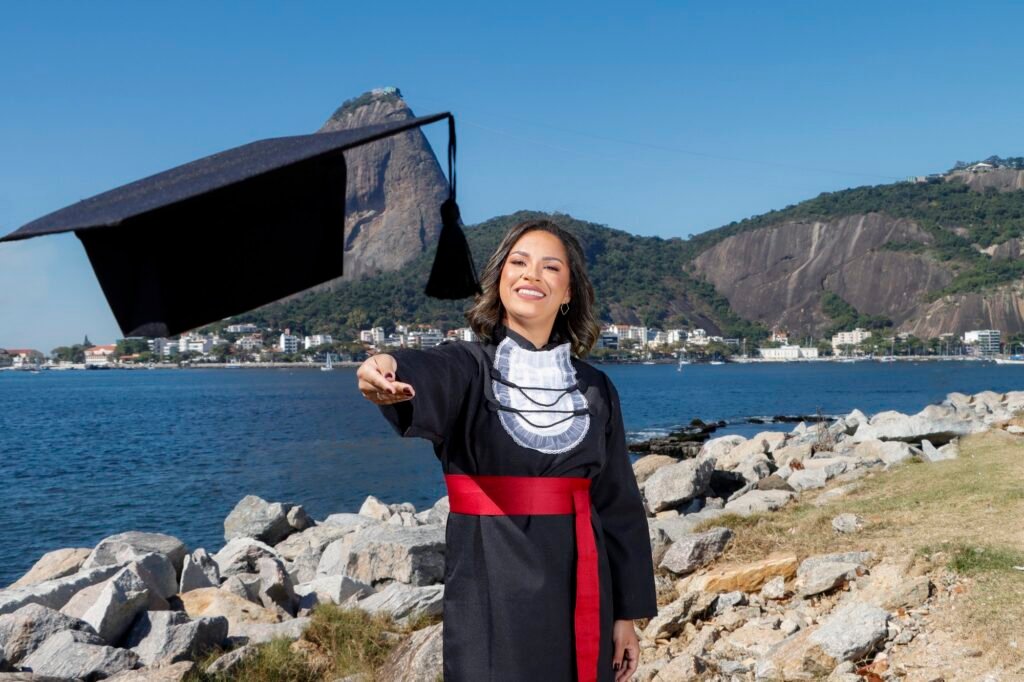 Formanda de Ciências Contábeis de faixa vermelha jogando o capelo à beira da baía, com o Pão de Açúcar ao fundo