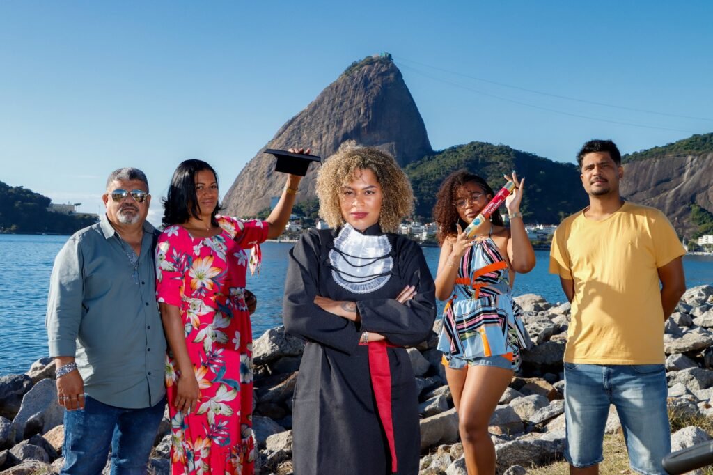 Formanda de Ciências Contábeis de braços cruzados com a família, que segura o capelo e o canudo, com o Pão de Açúcar ao fundo