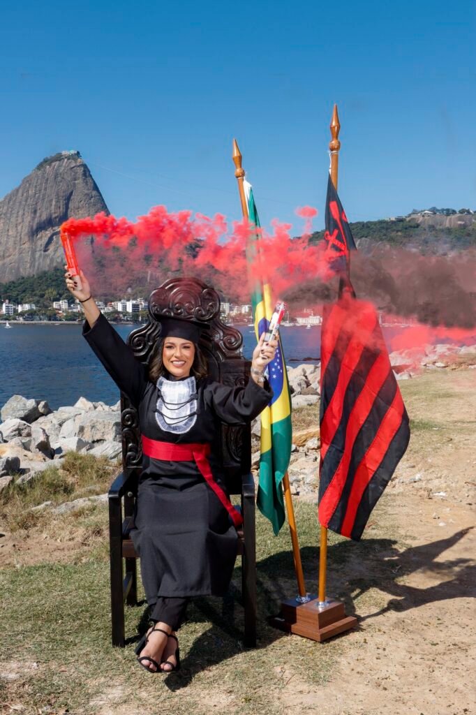 Formanda de Ciências Contábeis na Cadeira Colonial Luiz XV com sinalizadores de fumaça vermelha, entre a bandeira do Brasil e a do Flamengo, com o Pão de Açúcar ao fundo