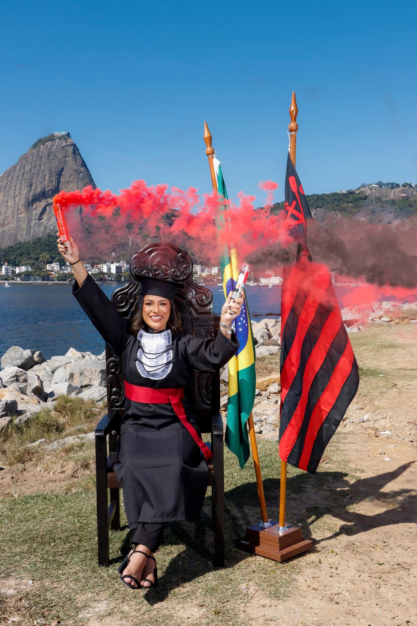 Formanda de Ciências Contábeis na Cadeira Colonial Luiz XV com sinalizadores de fumaça vermelha, entre a bandeira do Brasil e a do Flamengo, com o Pão de Açúcar ao fundo