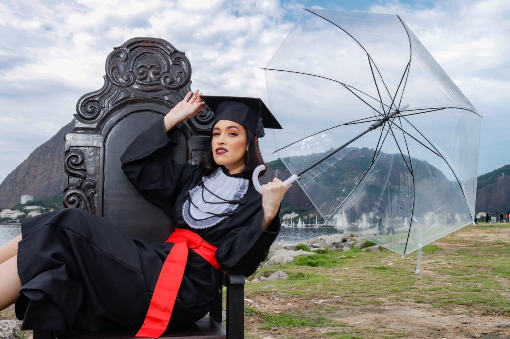Formanda de Direito de beca e faixa vermelha recostada na Cadeira Colonial Luiz XV com um guarda-chuva transparente, à beira da baía