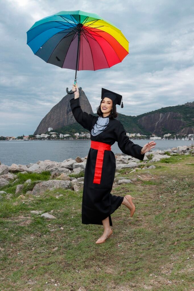Formanda de Direito com um guarda-chuva colorido, dando um passo de dança à beira da baía com o Pão de Açúcar ao fundo