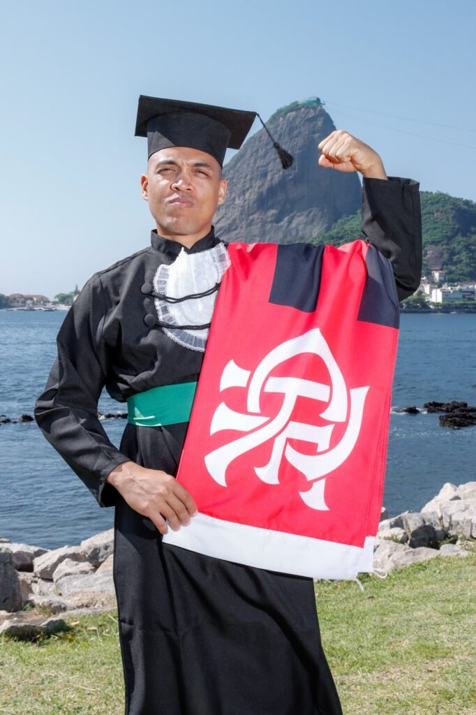 Formando de Educação Física com a bandeira do Flamengo no ombro, mostrando o bíceps, com o Pão de Açúcar ao fundo