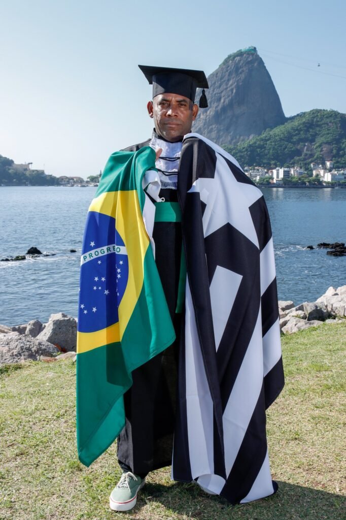 Formando de Educação Física de beca e capelo com a bandeira do Brasil e a do Botafogo nos ombros, com o Pão de Açúcar ao fundo