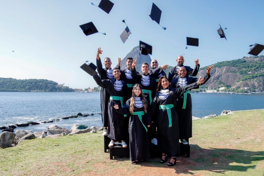 Turma de Educação Física de faixa verde jogando os capelos para o alto à beira da baía, com o Pão de Açúcar ao fundo