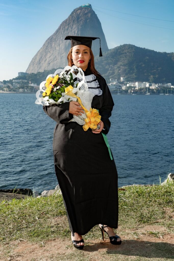 Formanda de Estética e Cosmetologia de beca e faixa verde com um buquê de girassóis, à beira da baía com o Pão de Açúcar