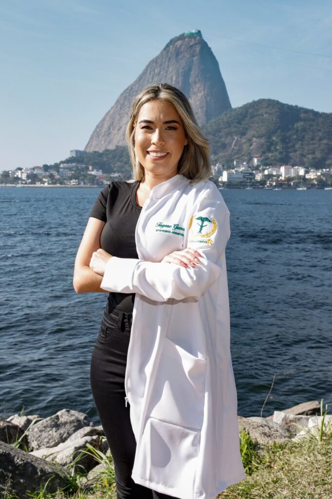 Formanda de Estética e Cosmetologia de braços cruzados, vestindo o jaleco bordado só de um lado, com o Pão de Açúcar ao fundo