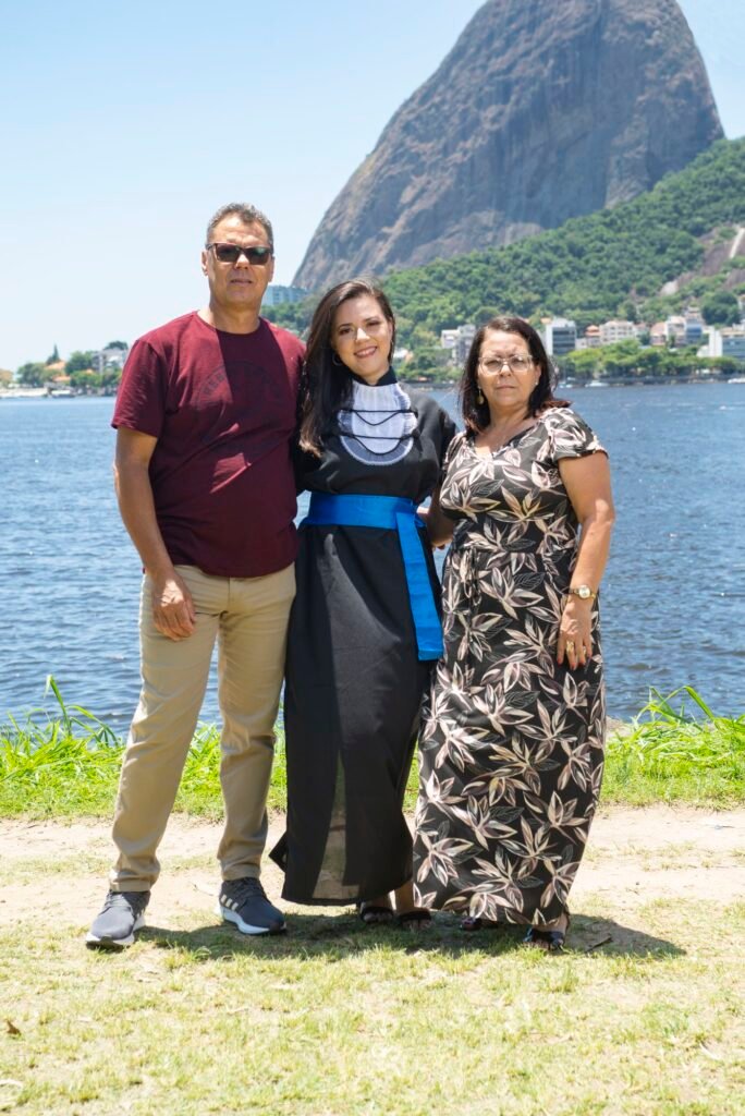 Formanda de beca e faixa azul ao lado dos pais à beira da baía, com o Pão de Açúcar ao fundo