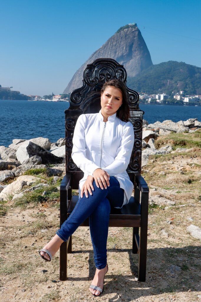 Formanda de jaleco branco e calça jeans sentada na Cadeira Colonial Luiz XV, com o Pão de Açúcar ao fundo