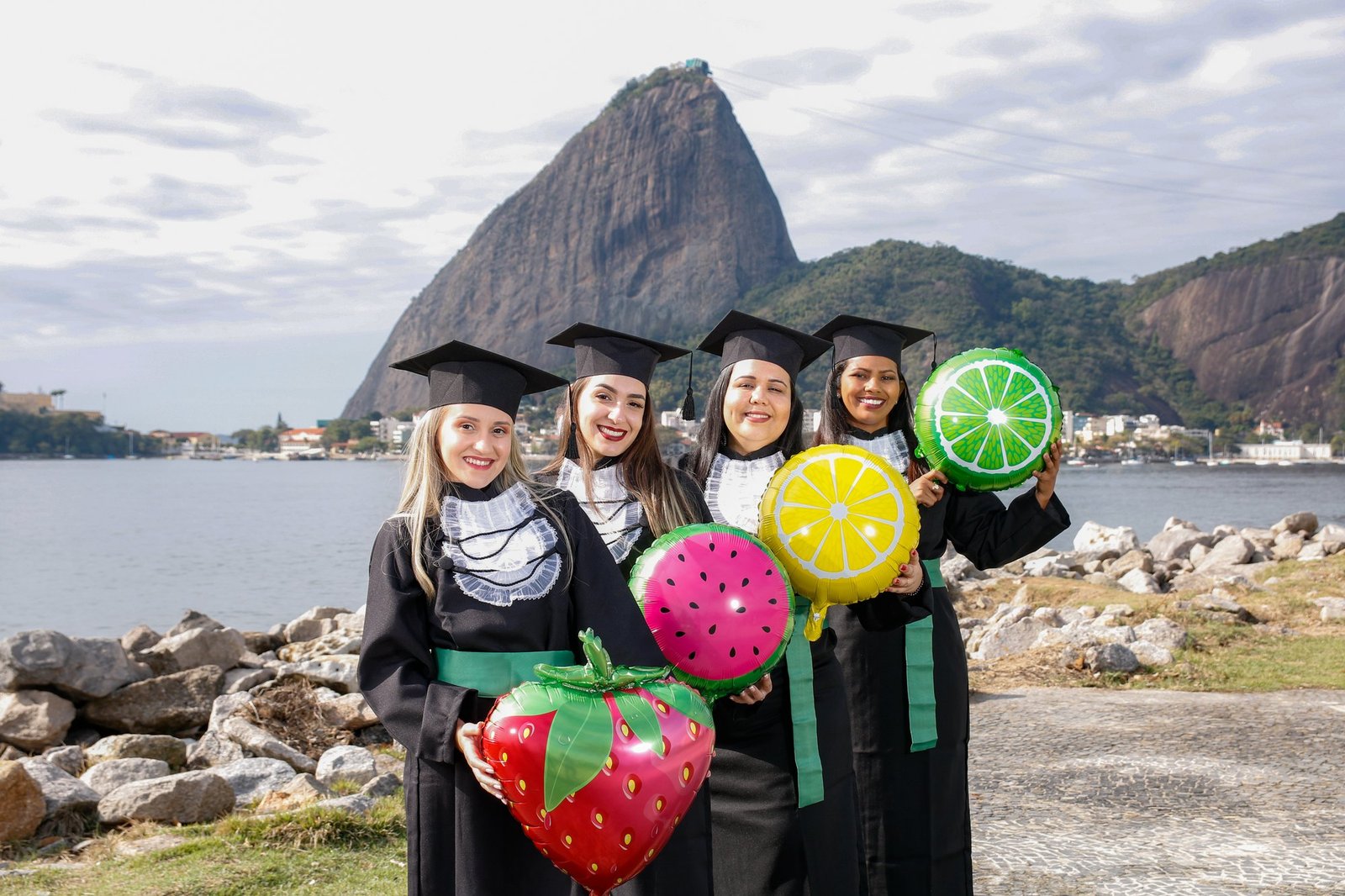 Quatro formandas de Nutrição de faixa verde segurando balões de morango, melancia, limão-siciliano e limão, com o Pão de Açúcar ao fundo