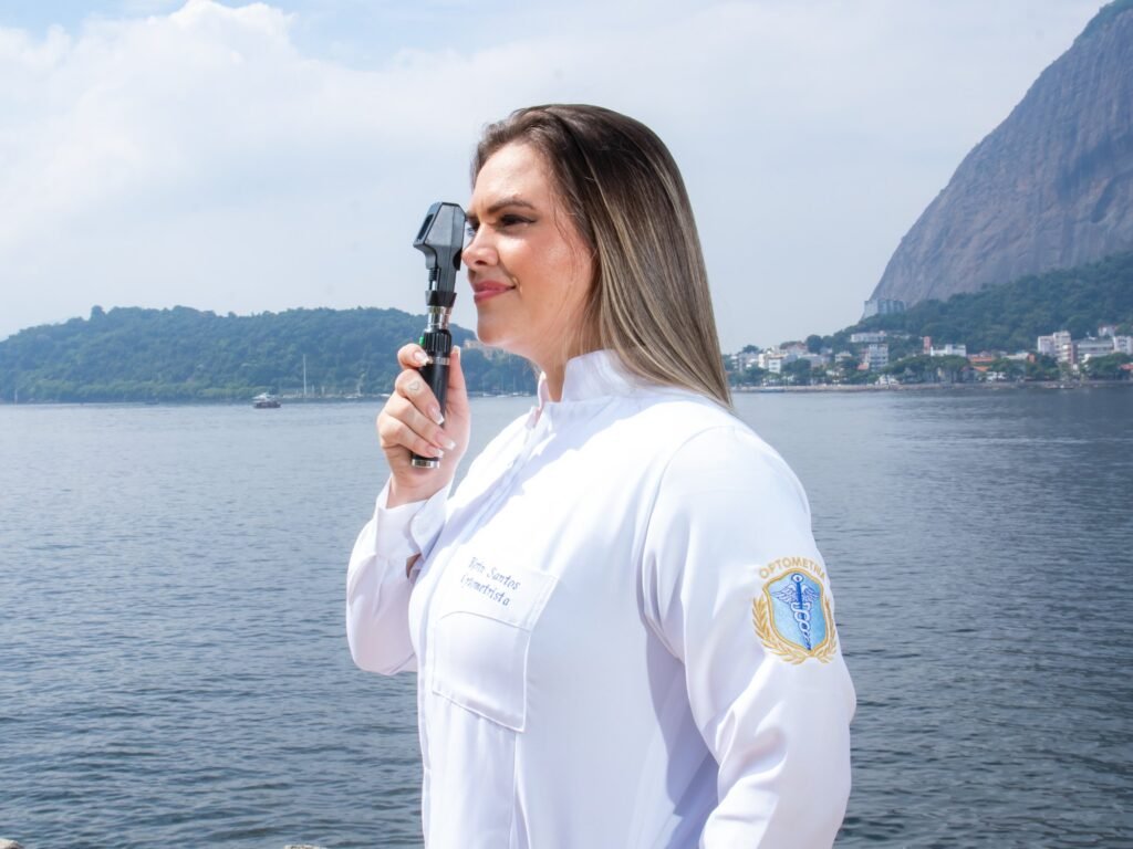 Formanda de Optometria de jaleco bordado segurando um oftalmoscópio junto ao rosto, à beira da baía com o Pão de Açúcar