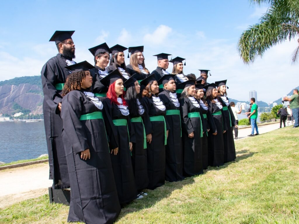 Turma de Optometria de beca e faixa verde em fileiras à beira da baía, num dia de céu azul