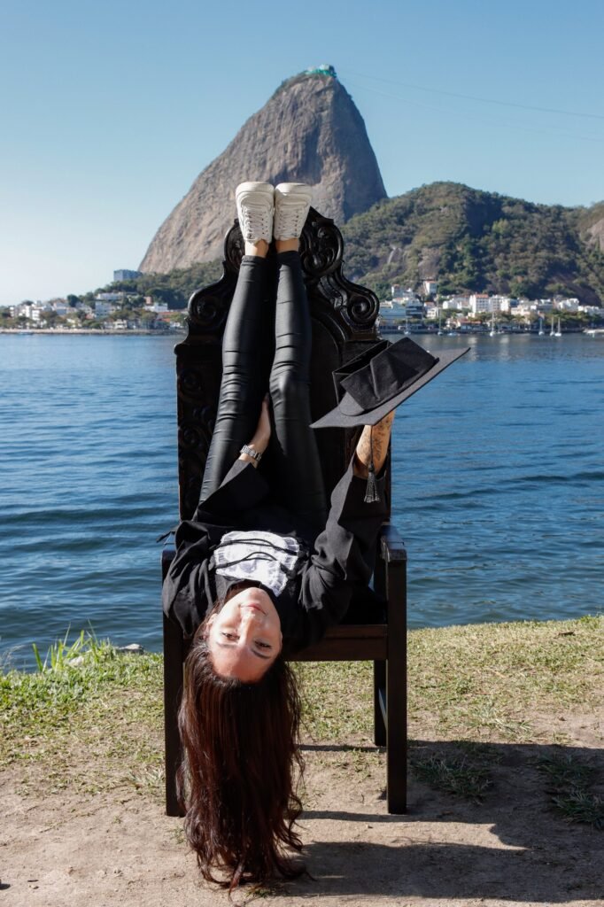 Formanda de Pedagogia de ponta-cabeça na Cadeira Colonial Luiz XV, segurando o capelo, com o Pão de Açúcar ao fundo