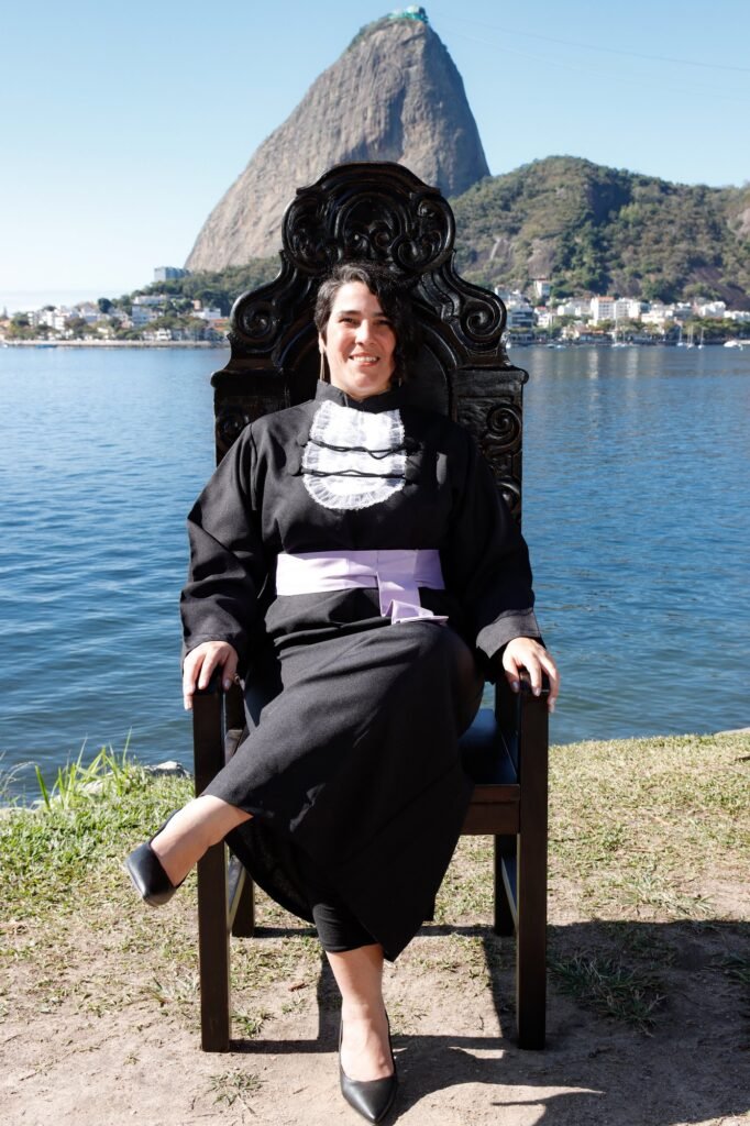 Formanda de Pedagogia de faixa lilás sentada na Cadeira Colonial Luiz XV à beira da baía, com o Pão de Açúcar ao fundo