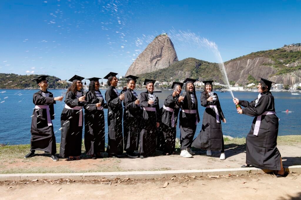 Turma de Pedagogia de beca e capelo brindando enquanto uma formanda estoura o espumante, com o Pão de Açúcar ao fundo
