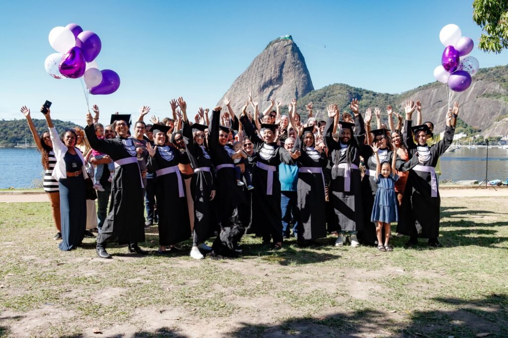 Turma de Pedagogia de faixa lilás com as famílias e balões roxos, de braços para o alto, com o Pão de Açúcar ao fundo