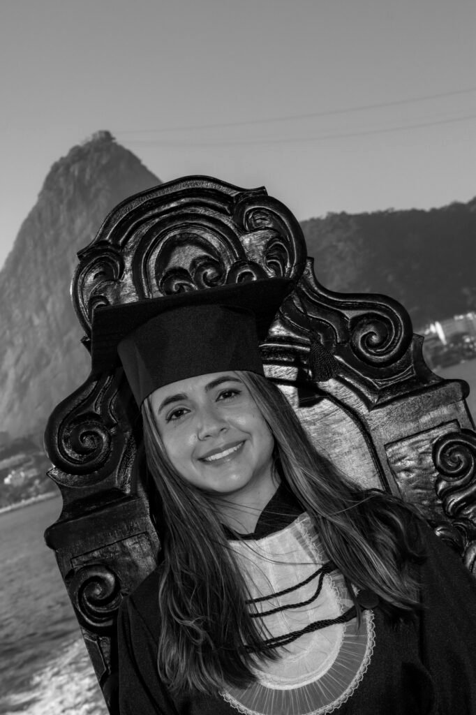 Formanda de Relações Internacionais de beca e capelo na Cadeira Colonial Luiz XV, em preto e branco, com o Pão de Açúcar ao fundo