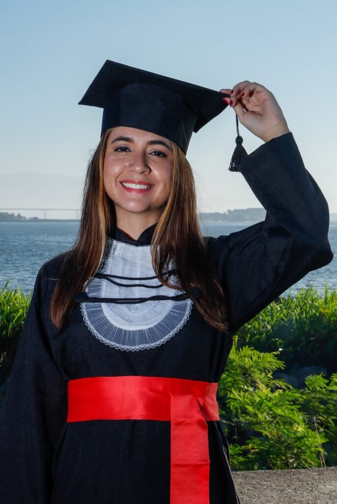 Formanda de Relações Internacionais de beca e faixa vermelha segurando o capelo, com a baía ao fundo