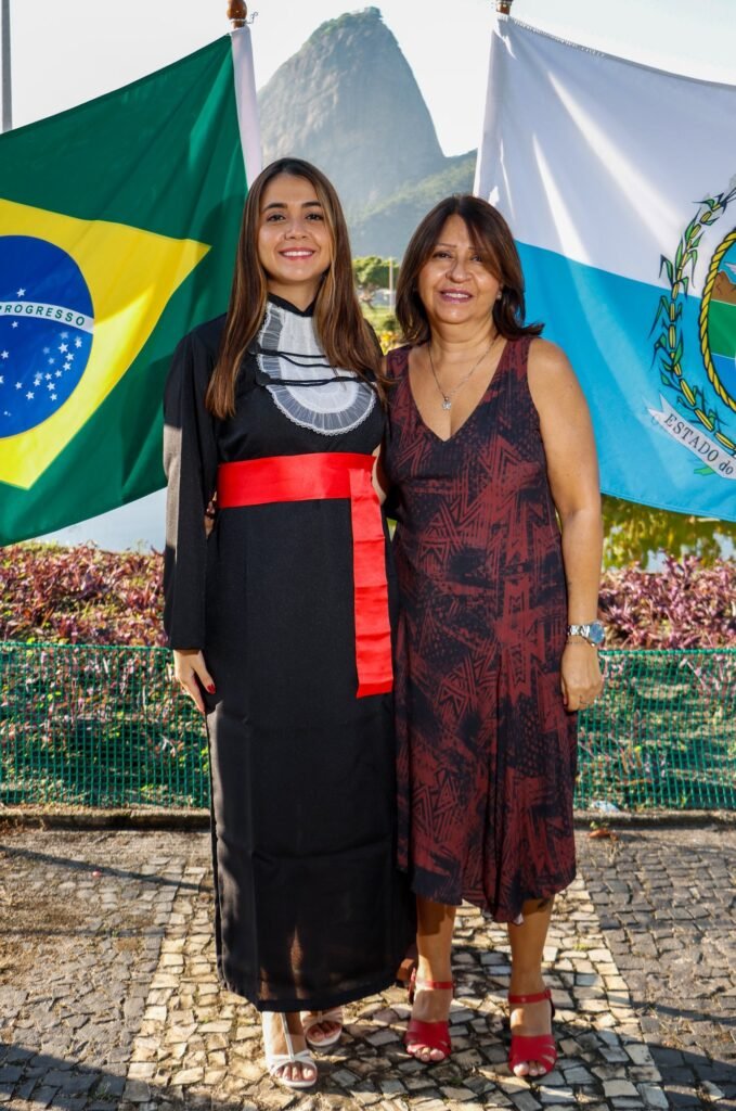 Formanda de Relações Internacionais com a mãe entre as bandeiras do Brasil e do Rio de Janeiro, no Monumento Estácio de Sá, com o Pão de Açúcar ao fundo