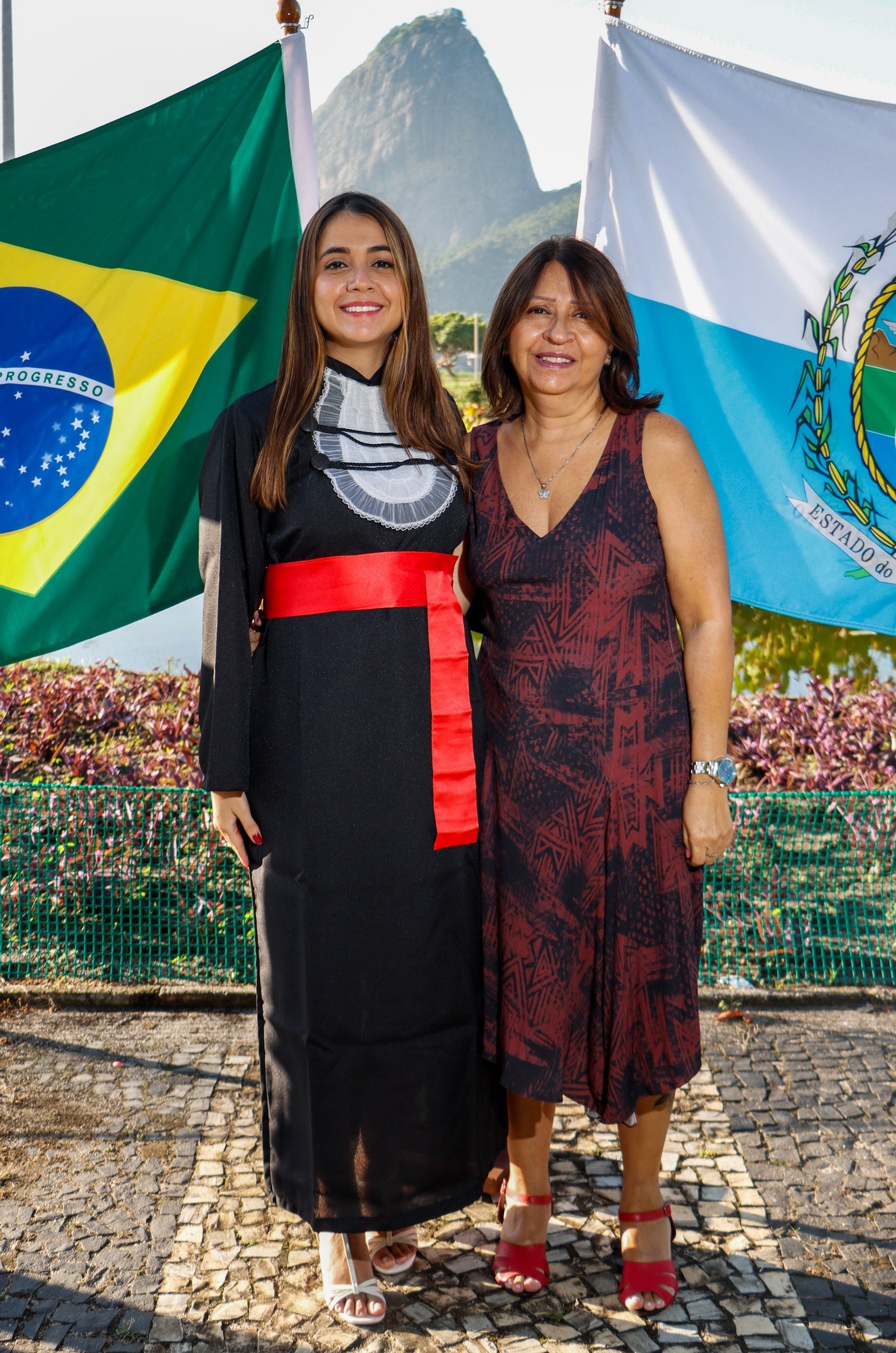 Formanda de Relações Internacionais com a mãe entre as bandeiras do Brasil e do Rio de Janeiro, no Monumento Estácio de Sá, com o Pão de Açúcar ao fundo