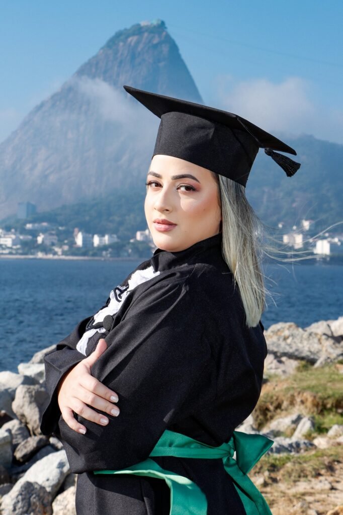 Formanda de beca e faixa verde de braços cruzados, olhando por cima do ombro, com o Pão de Açúcar coberto de nuvens ao fundo