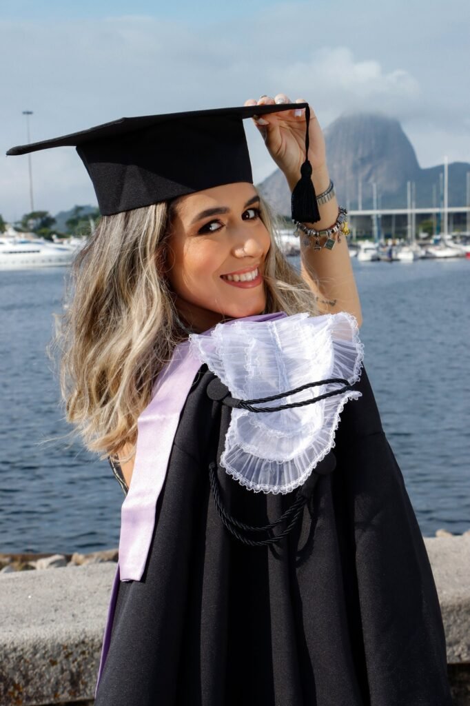 Formanda de Recursos Humanos de beca e faixa lilás olhando por cima do ombro e segurando o capelo, com barcos e o Pão de Açúcar ao fundo