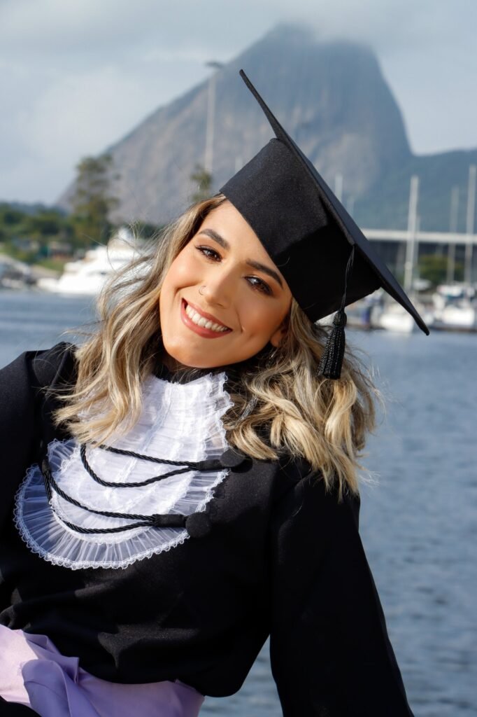 Formanda de Recursos Humanos sorrindo de capelo inclinado à beira da baía, com o Pão de Açúcar ao fundo