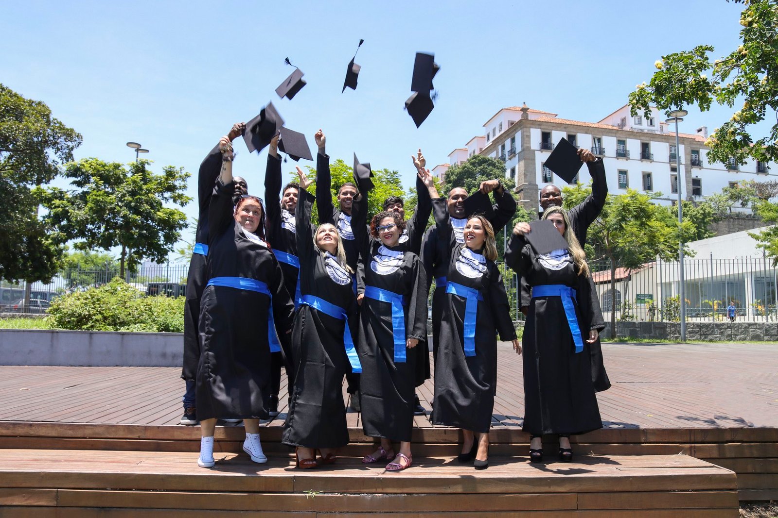 Turma de formandos de beca e faixa azul jogando o capelo para o alto em ensaio de formatura externo