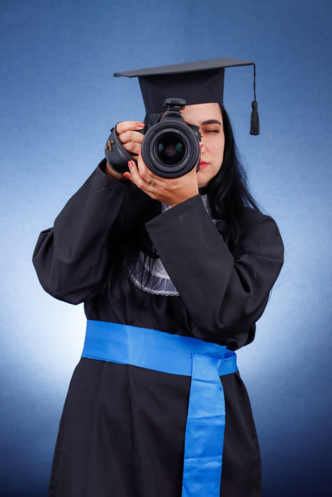 Formanda de Fotografia de beca e faixa azul fotografando com a câmera no fundo azul