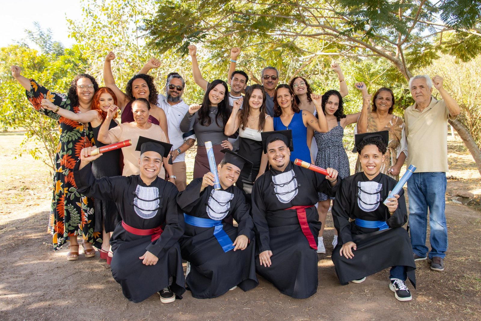 Quatro formandos de Ciências Contábeis, TI e Gestão agachados com os canudos, com as famílias comemorando atrás, no Parque do Gericinó