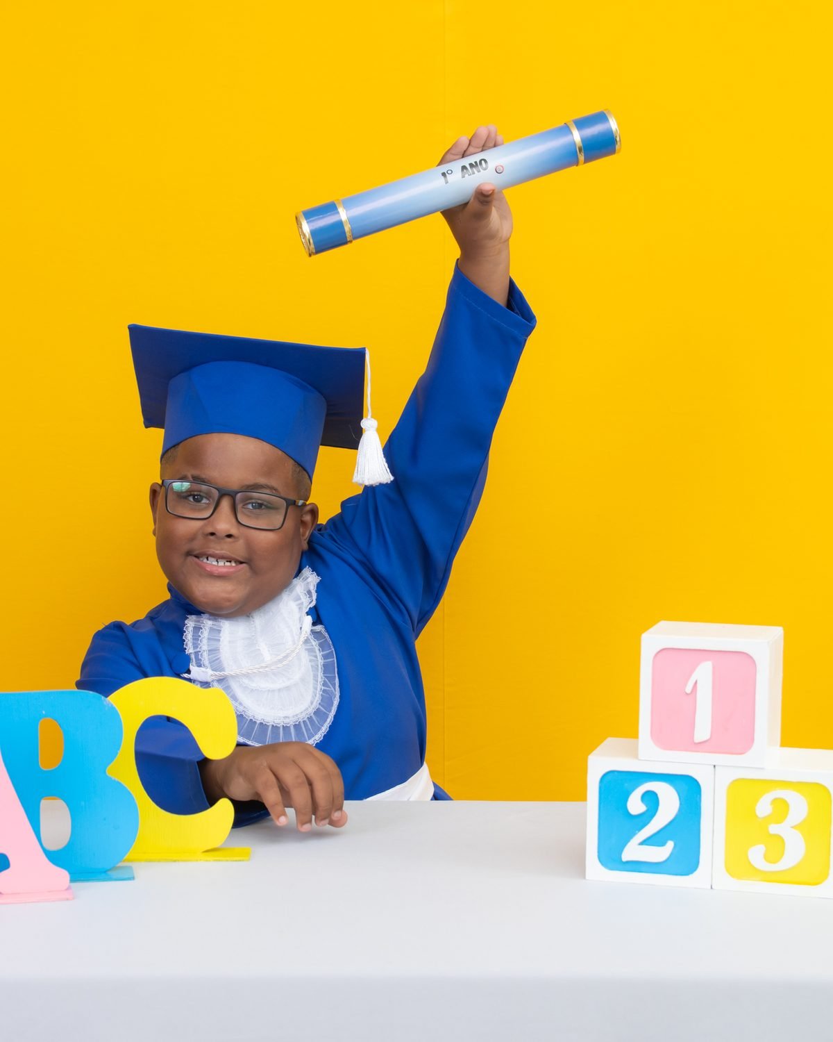 Menino de beca azul erguendo o canudo do 1º ano no cenário amarelo com letras ABC