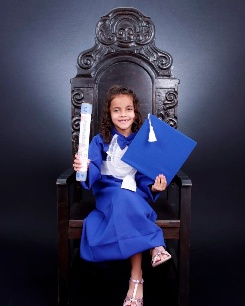 Menina de beca azul na cadeira entalhada com o canudo da Formatura ABC e o capelo